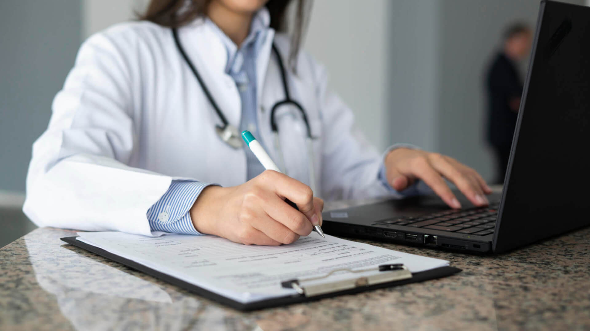 Doctor typing on laptop and writing on notepad.