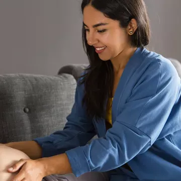 Foto einer schwangeren Frau, die auf einer Couch liegt und gerade von einer Hebamme untersucht wird